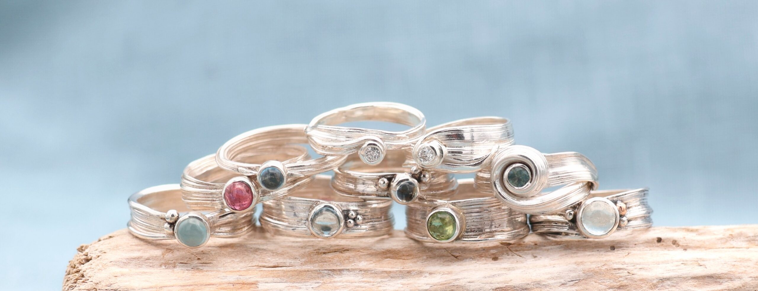 Photograph of rings with different coloured gems, designed and created by Marion, sitting on a piece of driftwood.