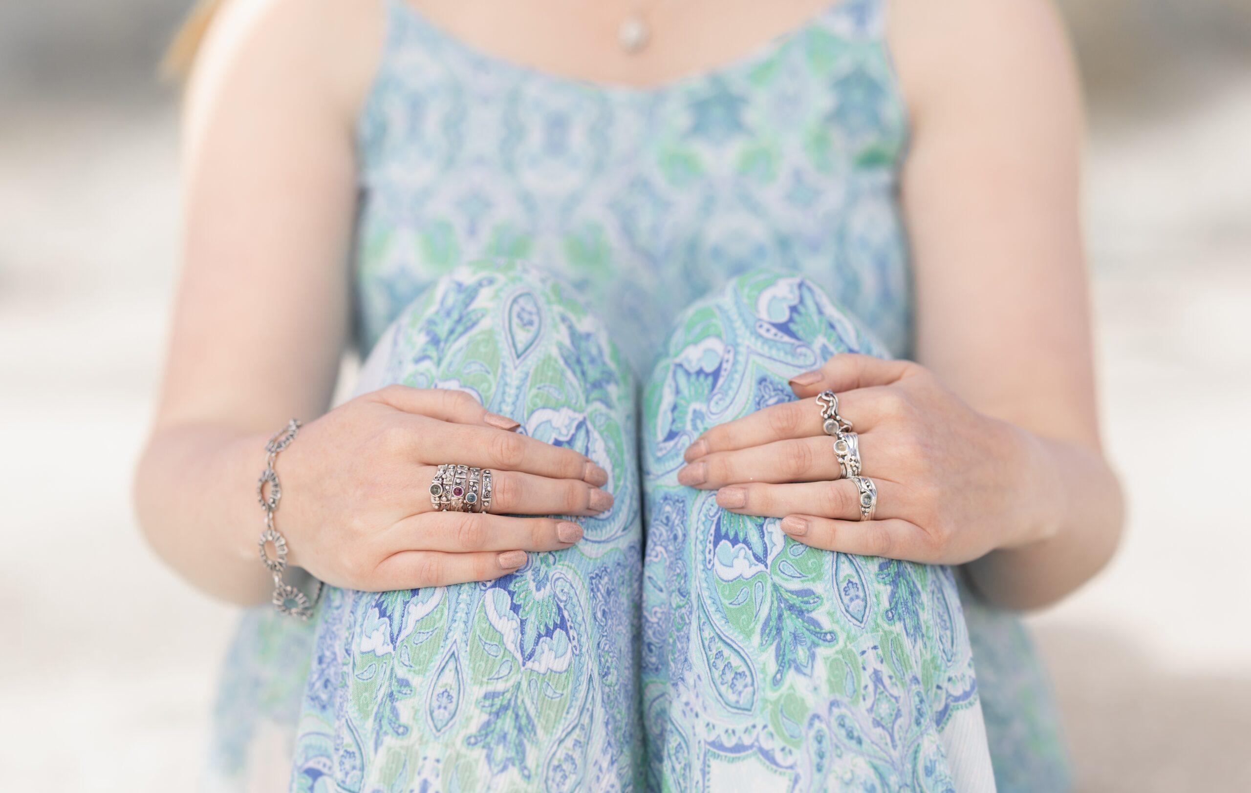 Photo of a woman sitting down, hugging her knees with the focus on her hands. She is wearing several rings designed by Marion.